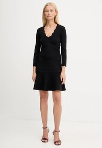 Donna in un top nero a maniche lunghe con bordi ondulati e una gonna a campana, che indossa sandali con tacco alto color bordeaux, in piedi contro uno sfondo bianco semplice.