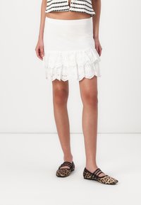 Jupe mini blanche avec un ourlet en couches et volants, présentant des détails en œillets. Associée à des chaussures plates en imprimé léopard avec des accents noirs.