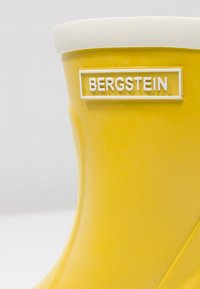 Geel rubberen regenlaars met een witte rubberen afwerking aan de bovenkant. Heeft een verhoogd "BERGSTEIN" logo in het wit aan de zijkant. Gladde textuur.
