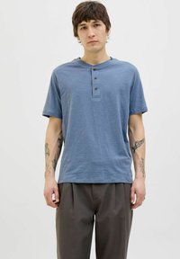 Kék, rövid ujjú henley póló három sötét gombbal és lekerekített nyakkal. Puha tapintású, laza szabású, sötét, redőzött nadrággal párosítva.