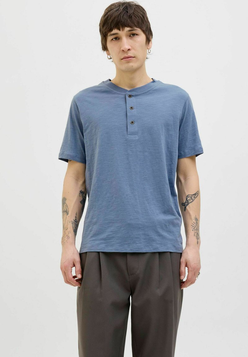 Kék, rövid ujjú henley póló három sötét gombbal és lekerekített nyakkal. Puha tapintású, laza szabású, sötét, redőzött nadrággal párosítva.