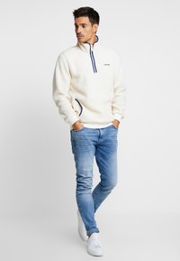Kridhvid fleece pullover med halv-zip og marineblå accenter; har en afslappet pasform og to sidelommer; parret med lyseblå jeans og hvide sko.