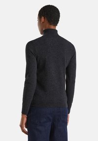 Personne portant un pull col roulé ajusté gris foncé et un jean bleu foncé, vu de dos sur un fond uni.