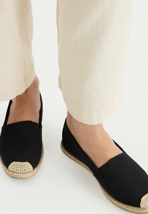 Füße in schwarzen Slip-On-Espadrilles mit beigen Zehenkappen und Jutsohlen, kombiniert mit hellbeigen, gekürzten Hosen.