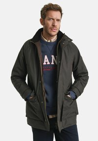 Donkergroene parka met rits- en drukknoopsluiting, voorzien van twee voorzakken en een hoge kraag, gelaagd over een marineblauwe sweatshirt.