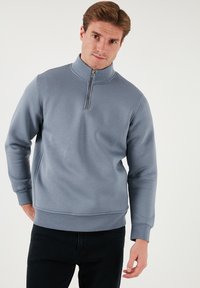 Felpa azzurra chiara con colletto alto e zip a metà, realizzata in materiale morbido. Presenta polsini a costine e una vestibilità comoda, abbinata a jeans scuri.