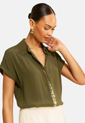 Žena oblečená v olivovo zelenej krátkorukávovej blúzke so zlatým flitrovaným lemom, krémovej sukni, zlatých kruhových náušniciach, ruka pri tvári, hľadí dole.