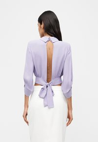 Γυναίκα με μακριά σκούρα μαλλιά φοράει μια λεβαντίν μπλούζα με ανοιχτή πλάτη, δεμένη στη μέση, και λευκή φούστα σε ένα απλό φόντο.