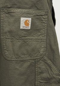 Τσέπη ελιάς παντελονιού Carhartt με ραμμένη λευκή ετικέτα που φέρει πορτοκαλί λογότυπο και όνομα μάρκας.