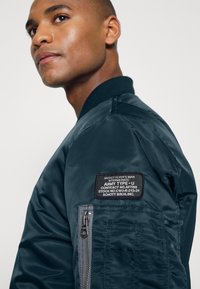 Giacca bomber navy con colletto lavorato a maglia, tasca con zip e etichetta in tessuto che dettaglia le specifiche militari. Texture liscia e lucida.
