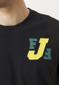 Short-sleeve black t-shirt with green and yellow block letters "FJ" and "E" printed on the front. Soft fabric, crew neckline, casual fit.