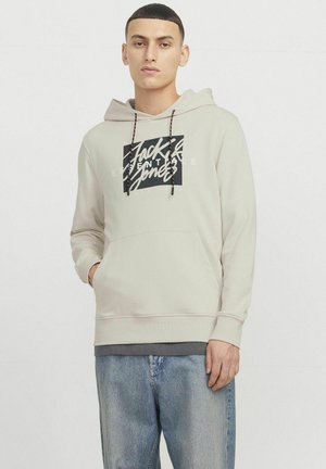 Lys beige hættejakke med snore, sort grafisk tekst foran, kængurulomme, ribbede manchetter, båret med lyseblå jeans.