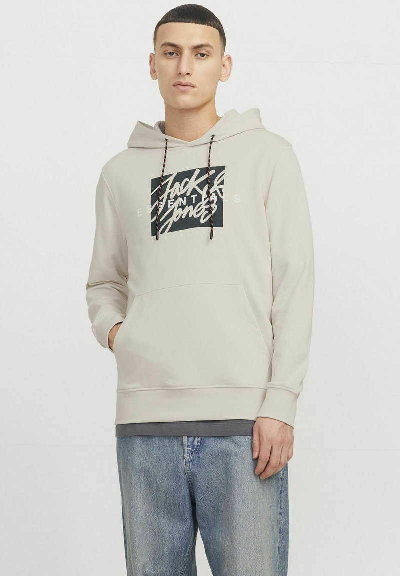Lys beige hættejakke med snore, sort grafisk tekst foran, kængurulomme, ribbede manchetter, båret med lyseblå jeans.