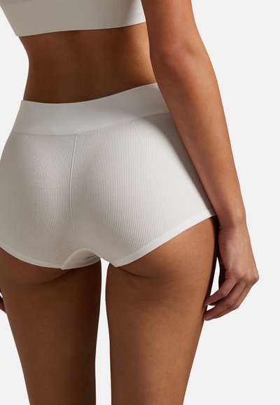 Kobieta w białych, prążkowanych bezszwowych szortach z wysokim stanem, pokazana od tyłu, w krótkim topie na jednolitym tle.