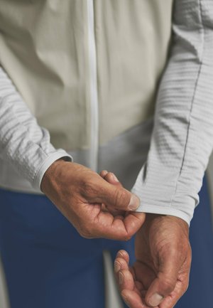 Person adjusting the cuff of a light gray, long-sleeve athletic shirt, wearing a beige vest and blue pants.
