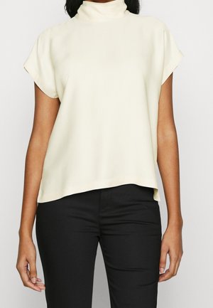 Vrouw draagt een crèmekleurige blouse met korte mouwen en een hoge hals, gecombineerd met zwarte strakke broek, staand tegen een effen achtergrond.