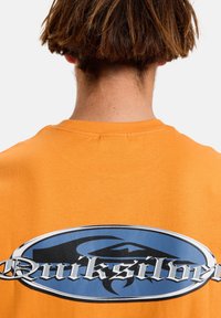 Oranžové bavlněné tričko s velkým modrým a stříbrným logem Quiksilver na zadní straně. Krátké rukávy a design s kulatým výstřihem.