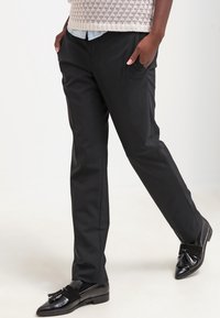 Pantaloni neri su misura con tasche frontali, design con fronte piatto e una texture liscia. Abbinati a mocassini neri lucidi con un dettaglio a fiocco.