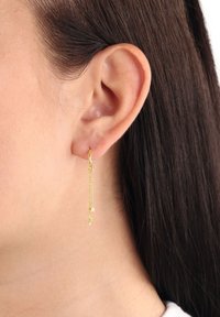 Kvinnas öra med mörkt hår som bär en delikat guldörhänge med två små stjärnformade berlocker hängande från en liten hoop.