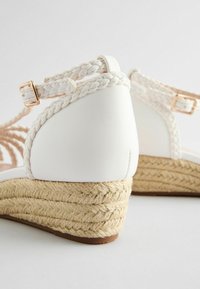 Bílé klínové sandály s pletenými popruhy a jute rope podrážkou. Disponují hladkým koženým vzhledem a zlatými přezkami.