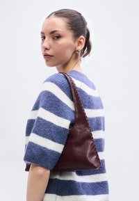 Geantă din piele burgundă cu margini împletite, având o formă triunghiulară. Modelul poartă un pulover în dungi albastre și albe, privirea îndreptată spre stânga.