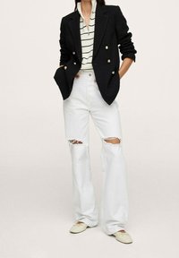 Kobieta w czarnym dwurzędowym żakiecie, w paskowanym swetrze na zamek, białych przetartych dżinsach i kremowych balerinach, z dłońmi w kieszeniach.