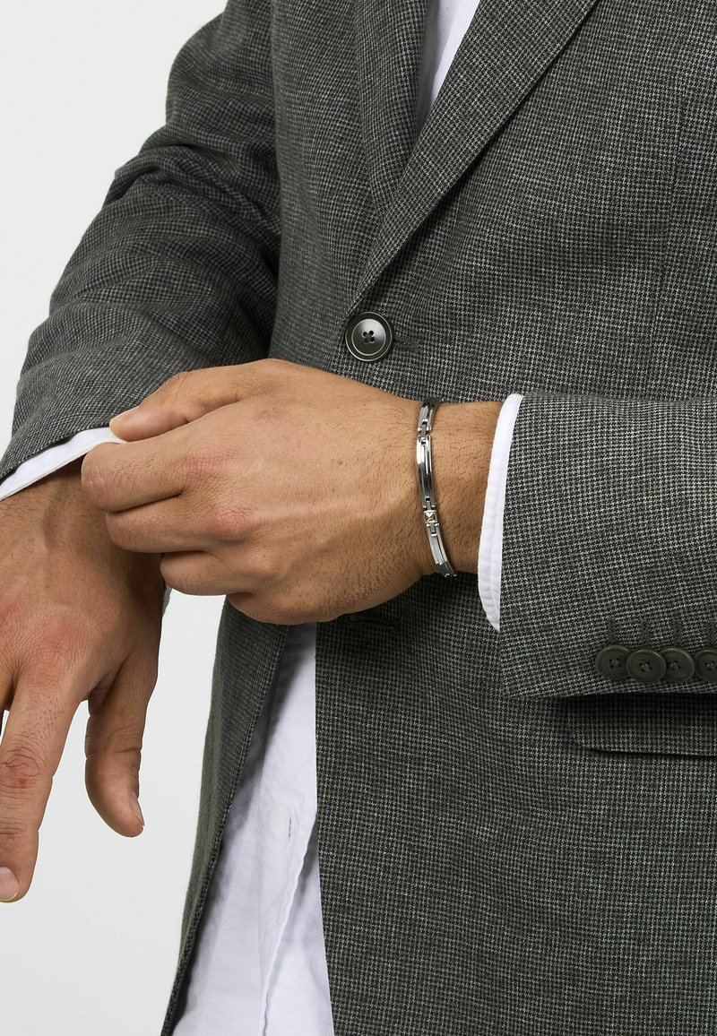 Blazer grigio a quadri con chiusura a due bottoni, tessuto leggermente strutturato e camicia bianca sotto; bracciale in metallo argentato al polso.