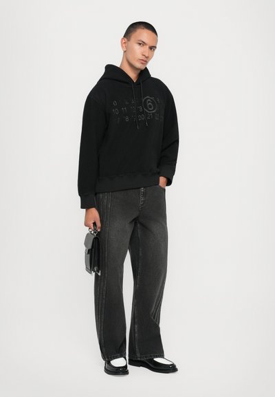 Svart hoodie med tonala tryckta siffror på framsidan, kombinerad med mörka, vida jeans med kontrasterande söm och svarta skor.