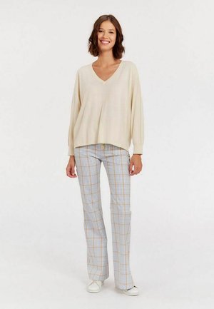 Pull crème en V avec une coupe ample et des manches longues, associé à un pantalon à carreaux bleu et jaune et des baskets blanches. Tissu texturé.