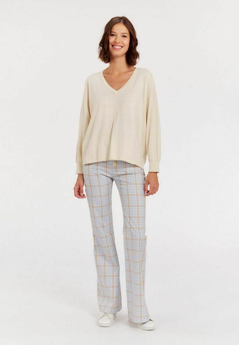 Pull crème en V avec une coupe ample et des manches longues, associé à un pantalon à carreaux bleu et jaune et des baskets blanches. Tissu texturé.