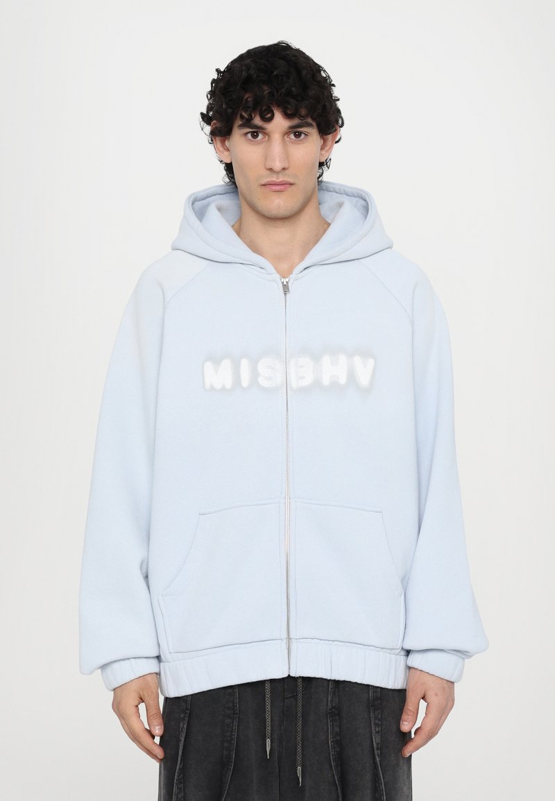 Lys blå hettejakke med fleece-struktur, store frontlommer, og "MISBHV" logo i hvit puffy skrift på brystet.