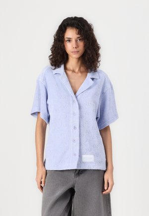 Chemise bleu clair à manches courtes, présentant un motif texturé, une fermeture à boutons et une petite étiquette à l'ourlet. Confectionnée en tissu doux.