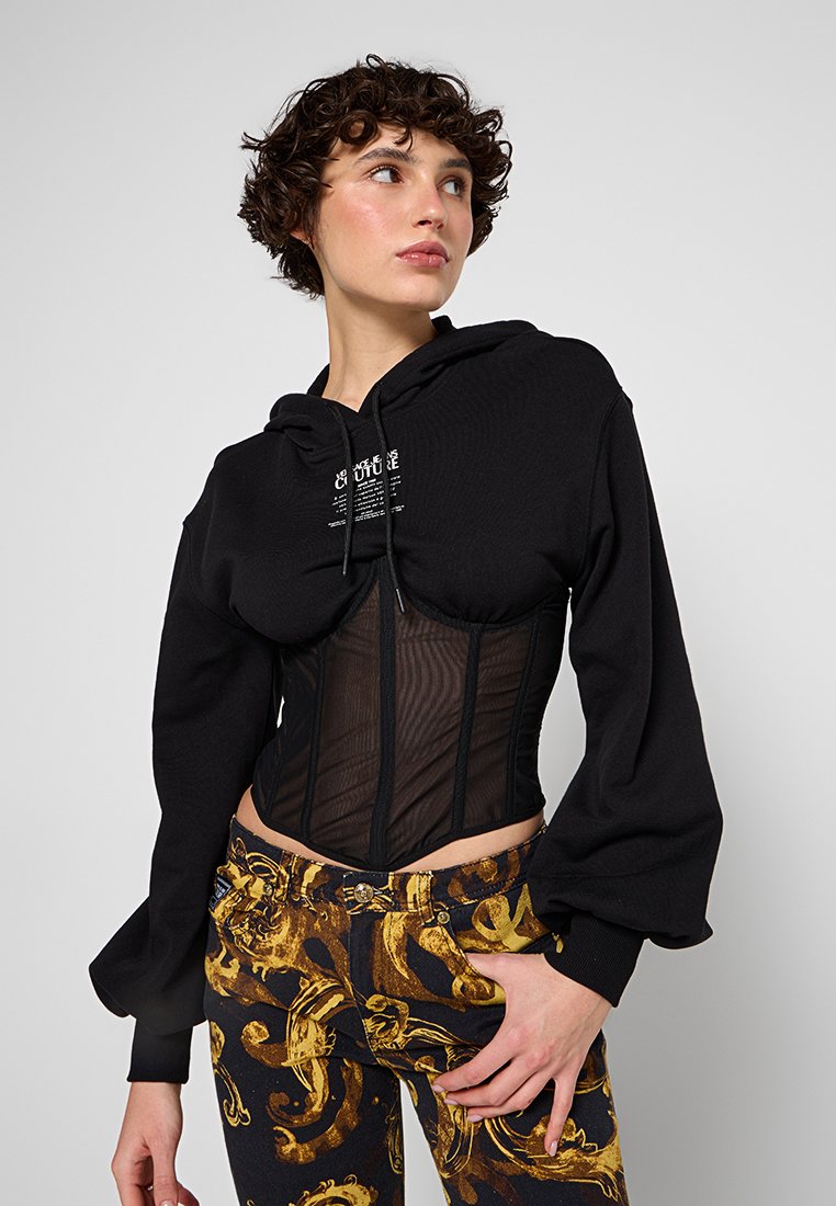 Sudadera negra con capucha y detalles de corsé transparente, con mangas largas tipo globo y un diseño ajustado. Combinada con pantalones estampados con detalles dorados.