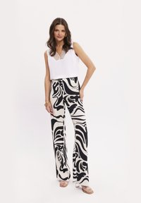 Biała, bezrękawnikowa bluzka z ozdobnym dekoltem, zestawiona z szerokimi spodniami w czarno-białe wzory i jasnymi sandałami.