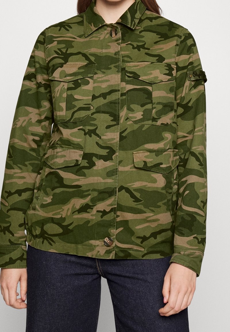 Femme portant une veste camouflage verte et marron avec des poches à rabat à boutons et un jean bleu foncé sur fond uni.