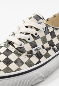 Baskets en toile avec un motif damier vert et crème, devant à lacets, œillets blancs et semelle en caoutchouc texturé. Logo "VANS" présent.