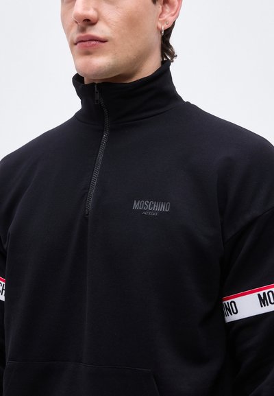 Άνδρας που φοράει μαύρο φούτερ Moschino Active με φερμουάρ στο ένα τέταρτο και λευκές και κόκκινες ρίγες με λογότυπο στα μανίκια, στέκεται μπροστά σε λευκό φόντο.