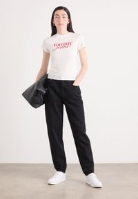 Valkoinen puuvillainen t-paita, jossa on punainen "tommy jeans" -logo, yhdistettynä mustiin housuihin ja mustaan nahkatakkiin, viimeisteltynä valkoisilla lenkkareilla.