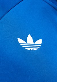 Λευκό λογότυπο Adidas trefoil κεντημένο σε φωτεινό μπλε ύφασμα, που δείχνει υφή πλεκτού υλικού και ραφές ρούχου.