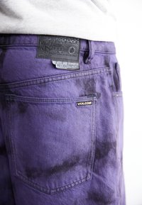 Pantalones cortos de mezclilla morados con un patrón de tie-dye, bolsillos delanteros y traseros, una etiqueta negra texturizada y bordado dorado de la marca en el costado.