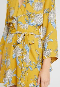 Florales Wickelkleid in Senfgelb mit blau-weißen Blumenmustern, gebundener Taille und weiten Ärmeln. Weicher, strukturierter Stoff.
