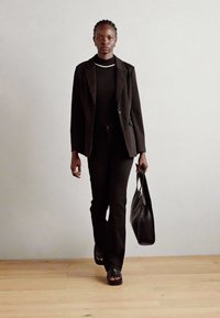 Zwarte blazer en getailleerde top, gecombineerd met zwarte uitlopende broek. Gladde leren tote bag en sandalen met open neus. Eenvoudig ontwerp, monotoon palet.