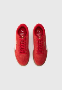 Røde ruskinds sneakers med et strømlinet design, der har en gummisål, røde snørebånd og subtile brandingdetaljer i guld og hvid.