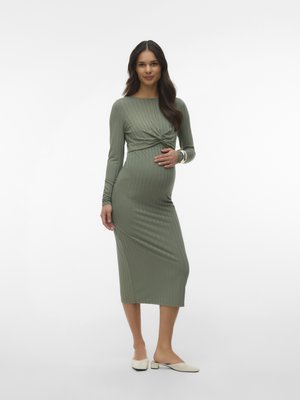 Femme enceinte aux longs cheveux foncés porte une robe midi côtelée à manches longues de couleur olive et des chaussures blanches à talons bas, debout sur un fond blanc.