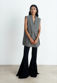 Nainen, jolla on pitkät mustat hiukset, pukeutuneena hihattomaan harmaaseen kaksiriviseen bleiseriin ja leveälahkeisiin mustiin housuihin, seisoo yksinkertaista seinää vasten.