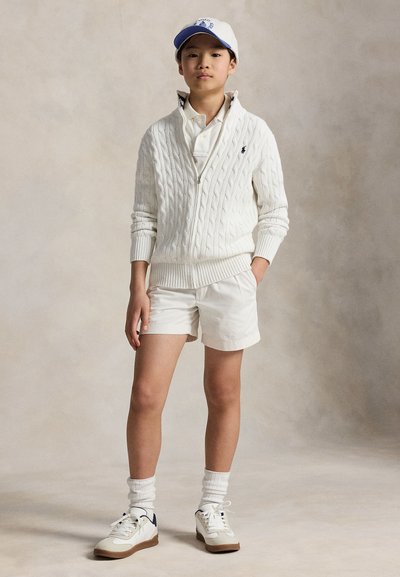 Junge Person steht und trägt einen weißen Kabelstrick-Reißverschluss-Pullover, weiße Shorts, weißes Poloshirt, weiße Socken, Turnschuhe und eine blau-weiße Baseballkappe.