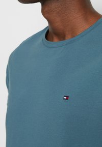 Tyrkysové bavlněné tričko s kulatým výstřihem, krátkými rukávy a malým vyšitým logem v červené, bílé a modré barvě.