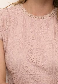 Top en dentelle rose tendre avec des motifs floraux et géométriques, encolure festonnée et manches courtes. Le superposé en dentelle texturée ajoute de la profondeur.