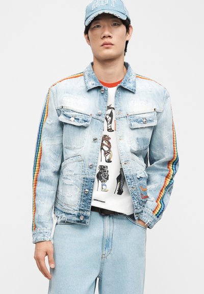 Νέος άνδρας που φοράει ανοιχτό μπλε τζιν μπουφάν με ρίγες ουράνιου τόξου στα μανίκια, λευκό μπλουζάκι με γραφικό σχέδιο, τζιν παντελόνι που ταιριάζει και ανοιχτό μπλε καπέλο.