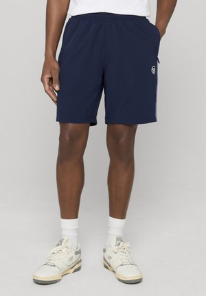 Mann iført marineblå treningsshorts, hvite sokker og hvite joggesko, står foran en nøytral bakgrunn.